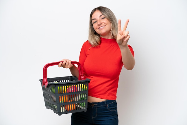 Foto jovem romena segurando uma cesta de compras cheia de comida isolada no fundo branco sorrindo e mostrando sinal de vitória