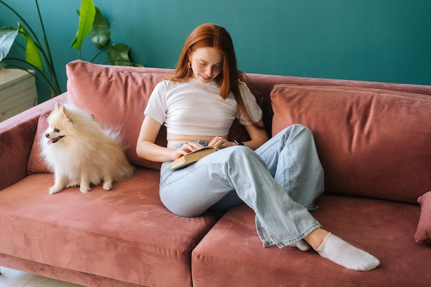 Jovem ruiva atraente lendo um livro sentado no sofá aconchegante com um cachorro spitz em miniatura