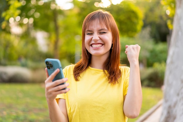 Jovem ruiva bonita ao ar livre usando telefone celular e fazendo gesto de vitória