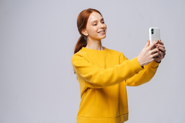 Jovem ruiva bonita assistindo a uma videochamada com o amante segurando um telefone inteligente na mão, tirando uma selfie