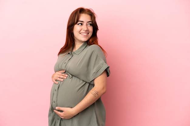 Jovem ruiva caucasiana isolada em um fundo rosa grávida e feliz