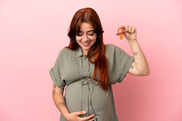 Jovem ruiva caucasiana isolada em um fundo rosa grávida e segurando uma chupeta