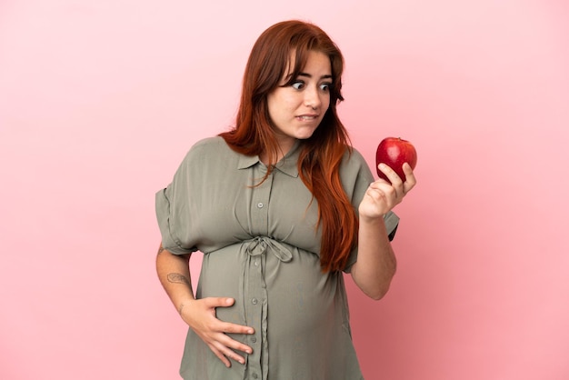 Jovem ruiva caucasiana isolada em um fundo rosa grávida e segurando uma maçã