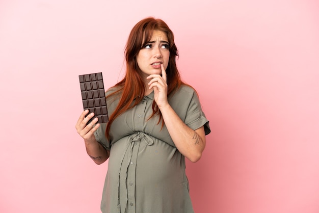 Jovem ruiva caucasiana isolada em um fundo rosa grávida e tendo dúvidas enquanto segura um chocolate