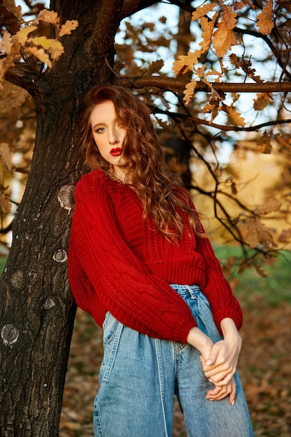 Jovem ruiva com um suéter vermelho caminha no parque. Retrato da beleza de outono de uma mulher ruiva elegante ao pôr do sol