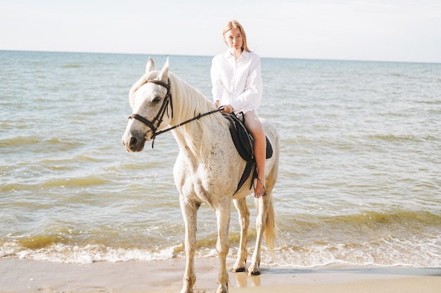 Jovem ruiva de camisa branca cavalgando cavalo branco no fundo da paisagem marinha