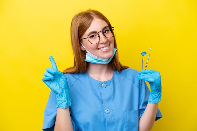 Jovem ruiva dentista isolada em fundo amarelo mostrando e levantando um dedo em sinal do melhor