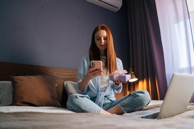 Jovem ruiva digitando mensagem no celular segurando a caixa de presente com presente. Fêmea bonita conversando no celular sentada na cama com as pernas cruzadas no quarto aconchegante luz perto do laptop.