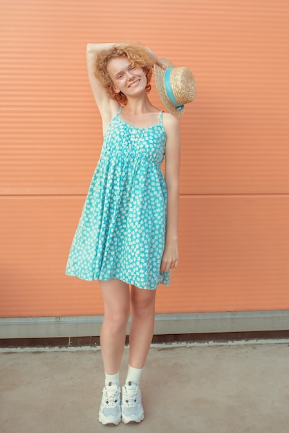 jovem ruiva encaracolada e alegre com um vestido de verão azul segurando um chapéu de palha em um background bege