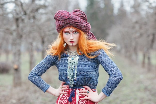 Jovem ruiva extravagante usando joias étnicas, roupas e turbante com maquiagem incomum, dançando ou posando em uma floresta mística ou parque. música trance psicodélica, ioga, conceito esotérico