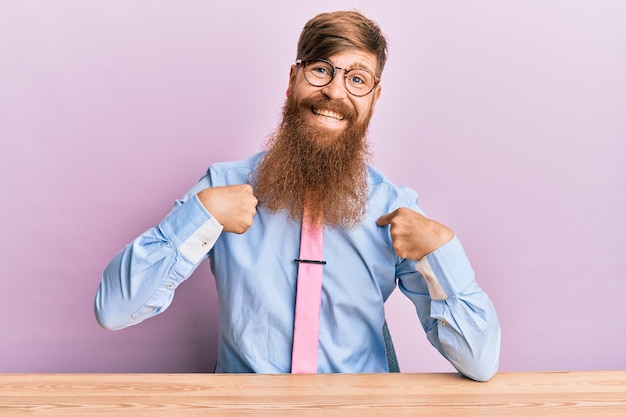 Jovem ruiva irlandesa vestindo camisa de negócios e gravata sentada na mesa parecendo confiante com um sorriso no rosto, apontando-se com os dedos orgulhosos e felizes.