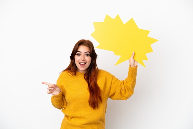 Jovem ruiva isolada em um fundo branco segurando um balão de fala vazio e apontando para o lado