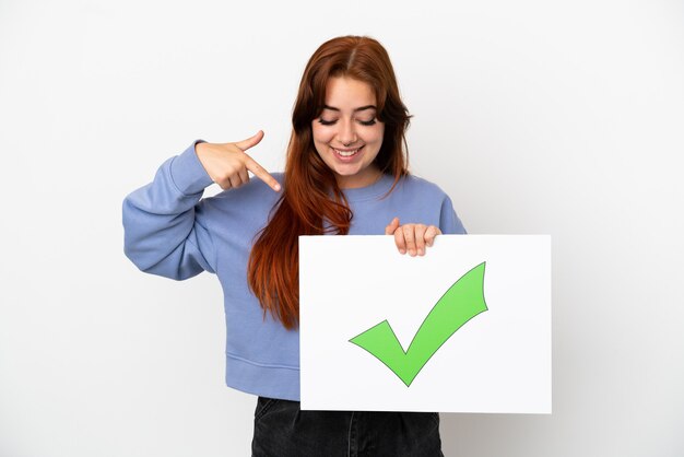 Jovem ruiva isolada em um fundo branco segurando um cartaz com o texto Ícone de marca de seleção verde e apontando-o