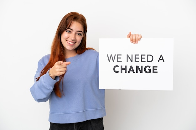 Jovem ruiva isolada em um fundo branco segurando um cartaz com o texto Precisamos de uma mudança e apontando para a frente