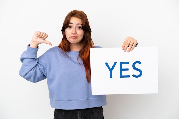 Jovem ruiva isolada em um fundo branco segurando um cartaz com o texto SIM com um gesto orgulhoso