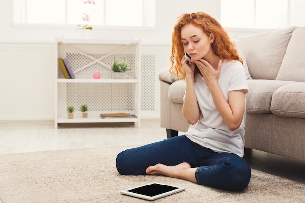 Jovem ruiva ligando para o celular enquanto usa o tablet digital, sentada no sofá bege de casa