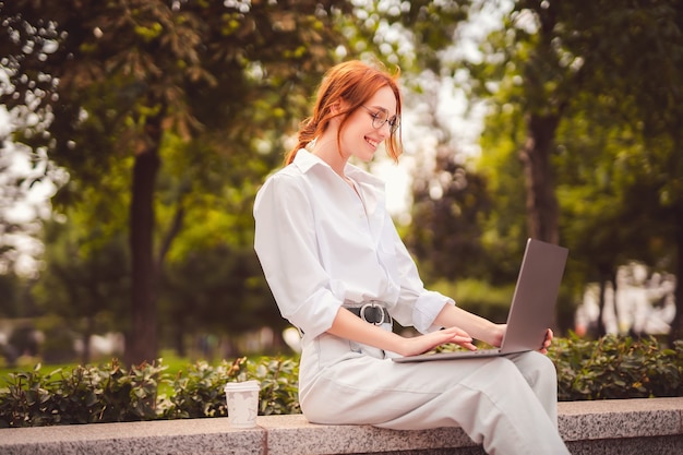 Jovem ruiva linda sentada no parque e usando laptop estudante universidade freelance wear ...