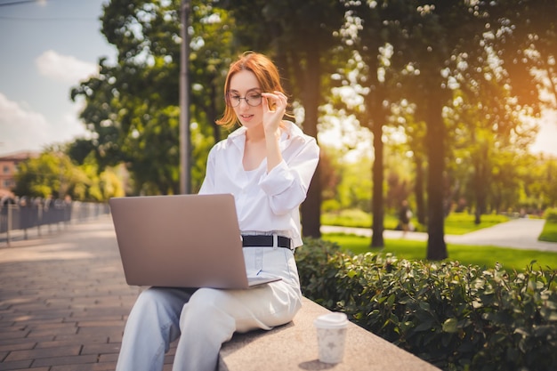 Jovem ruiva linda sentada no parque e usando laptop estudante universidade freelance wear ...