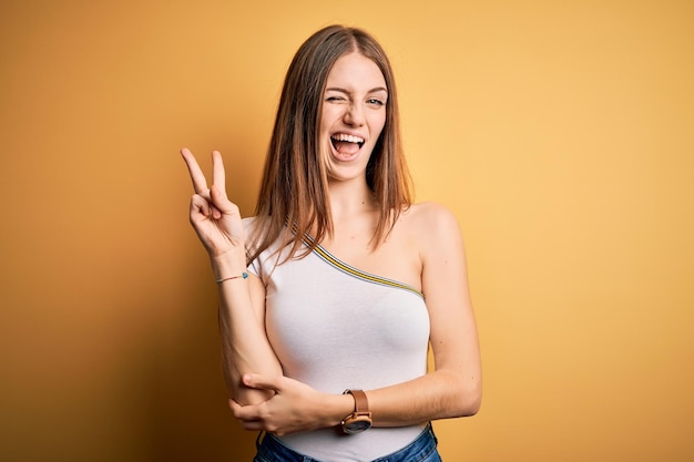 Jovem ruiva linda vestindo camiseta casual sobre fundo amarelo isolado, sorrindo com cara feliz, piscando para a câmera, fazendo sinal de vitória com os dedos número dois