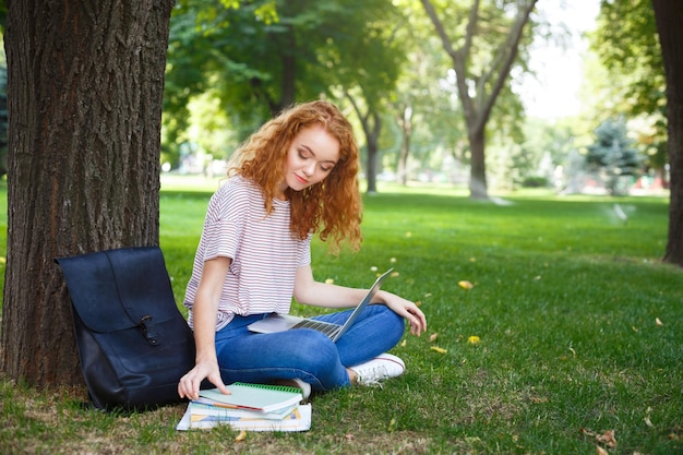 Jovem ruiva olhando para os blocos de notas, segurando o laptop no joelho, sentado com as pernas cruzadas sob uma árvore no parque. Conceito de estilo de vida educacional