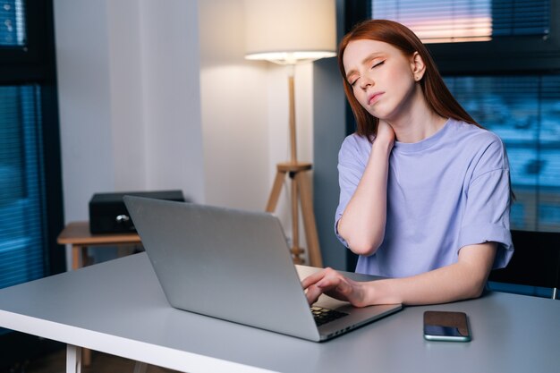 Jovem ruiva sobrecarregada digitando no laptop ativamente sentindo dor no pulso, sentado na mesa perto da janela, à noite. senhora trabalhando no laptop e sofrendo de dores nas mãos, síndrome do túnel do carpo.