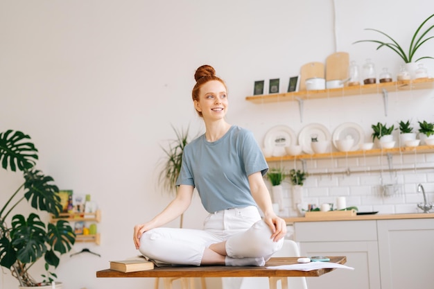 Jovem ruiva sorridente sentada em pose de lótus na mesa e olhando para longe garota feliz vestindo roupas casuais brancas meditando no escritório em casa conceito de descanso durante o trabalho remoto em auto-isolamento