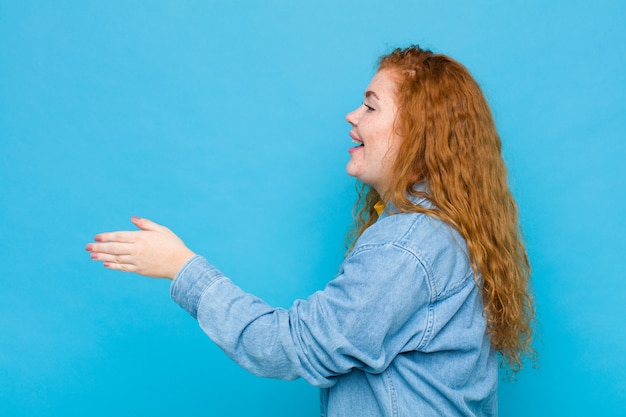 Jovem ruiva sorrindo, cumprimentando você e oferecendo um aperto de mão para fechar um negócio de sucesso, conceito de cooperação contra a parede azul