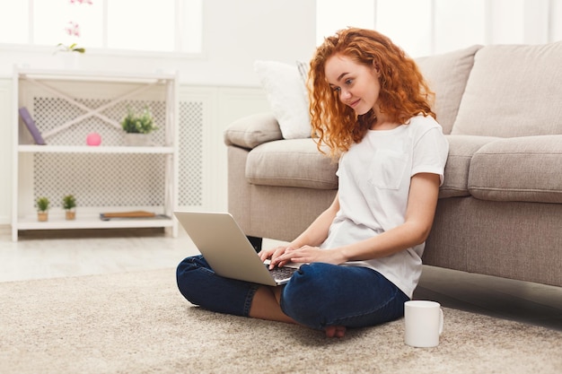 Jovem ruiva trabalhando no laptop sentada no chão com café