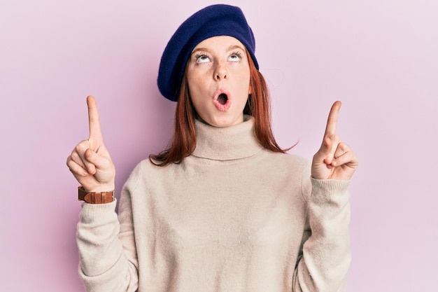 Jovem ruiva vestindo look francês com boina espantada e surpresa olhando para cima e apontando com os dedos e braços erguidos.