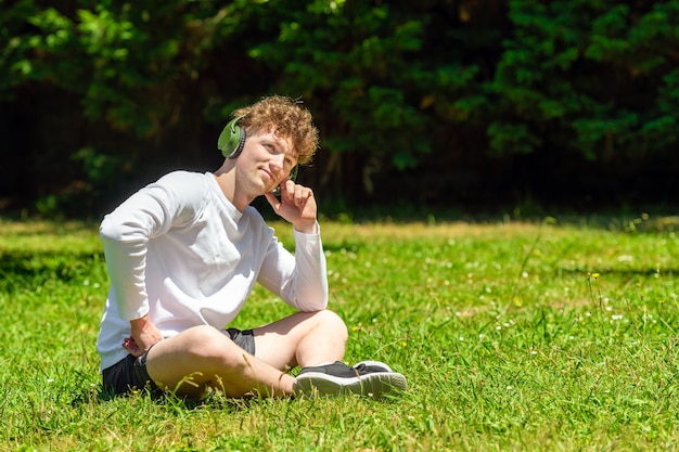 Foto jovem ruivo com fones de ouvido sentado na grama verde em um dia ensolarado