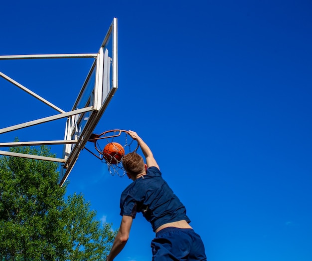 Jovem ruivo com uma camiseta azul escura joga uma bola em movimento em uma cesta de basquete contra um céu azul ao ar livre