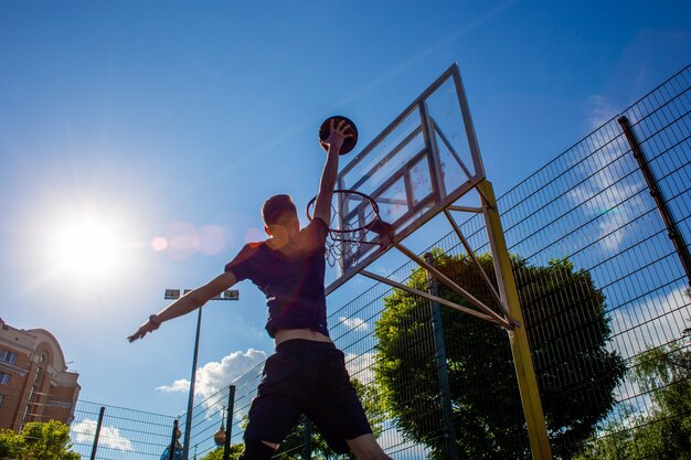 Jovem ruivo com uma camiseta azul escura joga uma bola em movimento em uma cesta de basquete contra um céu azul ao ar livre