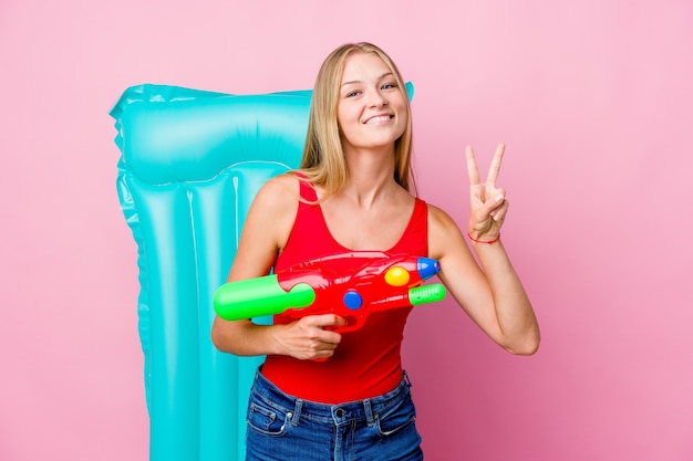 Jovem russa brincando com uma pistola de água com um colchão de ar, mostrando o sinal de vitória e sorrindo amplamente.