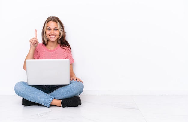 Jovem russa com um laptop sentado no chão isolado em fundo branco mostrando e levantando um dedo em sinal do melhor