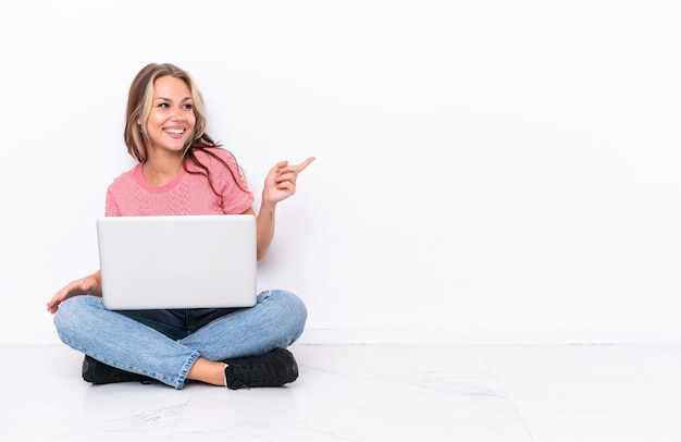 Jovem russa com um laptop sentado no chão isolado no fundo branco apontando o dedo para o lado e apresentando um produto