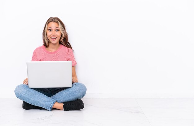 Jovem russa com um laptop sentado no chão isolado no fundo branco com expressão facial de surpresa