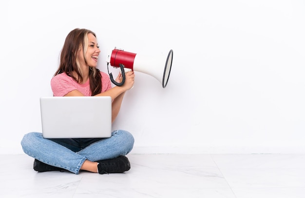 Jovem russa com um laptop sentado no chão isolado no fundo branco gritando através de um megafone