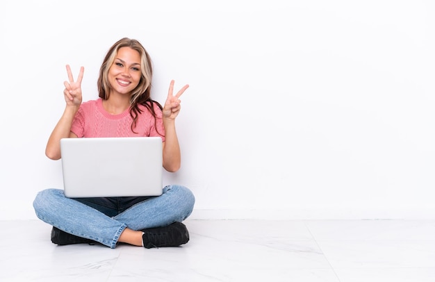 Jovem russa com um laptop sentado no chão isolado no fundo branco, mostrando sinal de vitória com as duas mãos