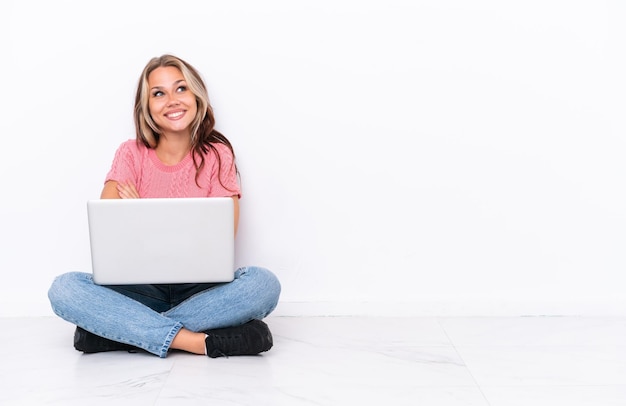 Jovem russa com um laptop sentado no chão isolado no fundo branco olhando para cima enquanto sorria