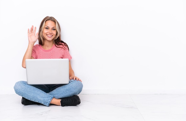 Jovem russa com um laptop sentado no chão isolado no fundo branco saudando com a mão com expressão feliz