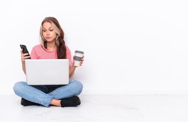 Jovem russa com um laptop sentado no chão isolado no fundo branco segurando café para levar e um celular