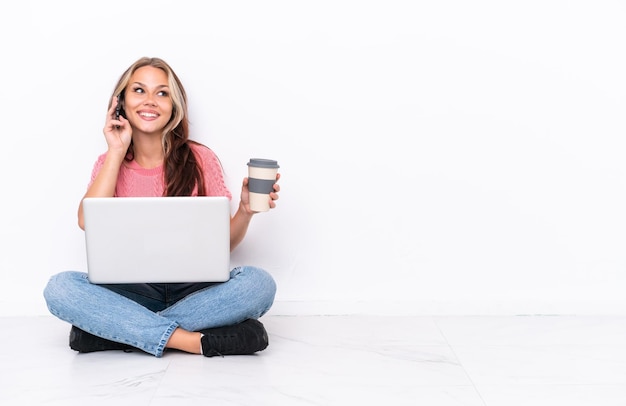 Jovem russa com um laptop sentado no chão isolado no fundo branco segurando café para levar e um celular