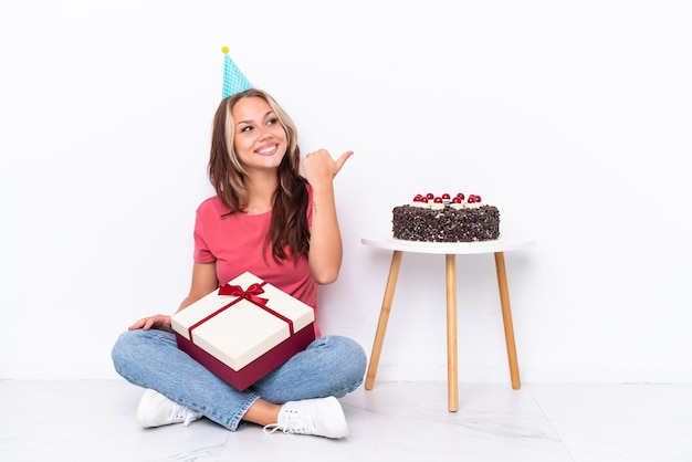 Jovem russa comemorando um aniversário sentada no chão isolada no fundo branco apontando para o lado para apresentar um produto