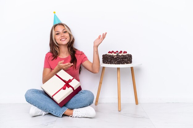 Jovem russa comemorando um aniversário sentada no chão isolada no fundo branco, estendendo as mãos para o lado para convidar para vir