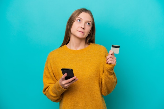 Jovem russa isolada em fundo azul comprando com o celular com cartão de crédito enquanto pensava