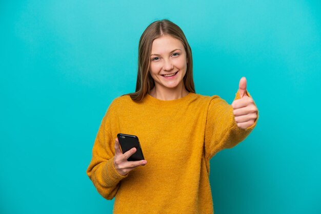 Jovem russa isolada em fundo azul usando telefone celular enquanto faz os polegares para cima
