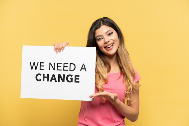 Jovem russa isolada em um fundo amarelo segurando um cartaz com o texto Precisamos de uma mudança e apontando-o