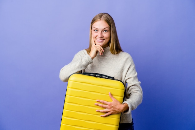 Jovem russa segurando a mala para viajar sorrindo feliz e confiante, tocando o queixo com a mão.