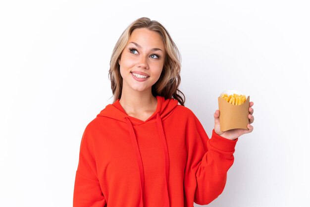 Jovem russa segurando batatas fritas isoladas em fundo branco pensando em uma ideia enquanto olha para cima