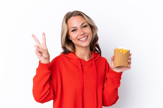 Jovem russa segurando batatas fritas isoladas no fundo branco, sorrindo e mostrando o sinal da vitória